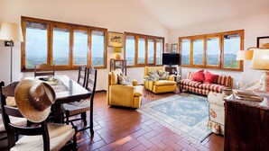 A spacious living room with a wooden dining table, yellow sofas, and a large window offering a view of the countryside. - Villa Pietraia (Certaldo)
