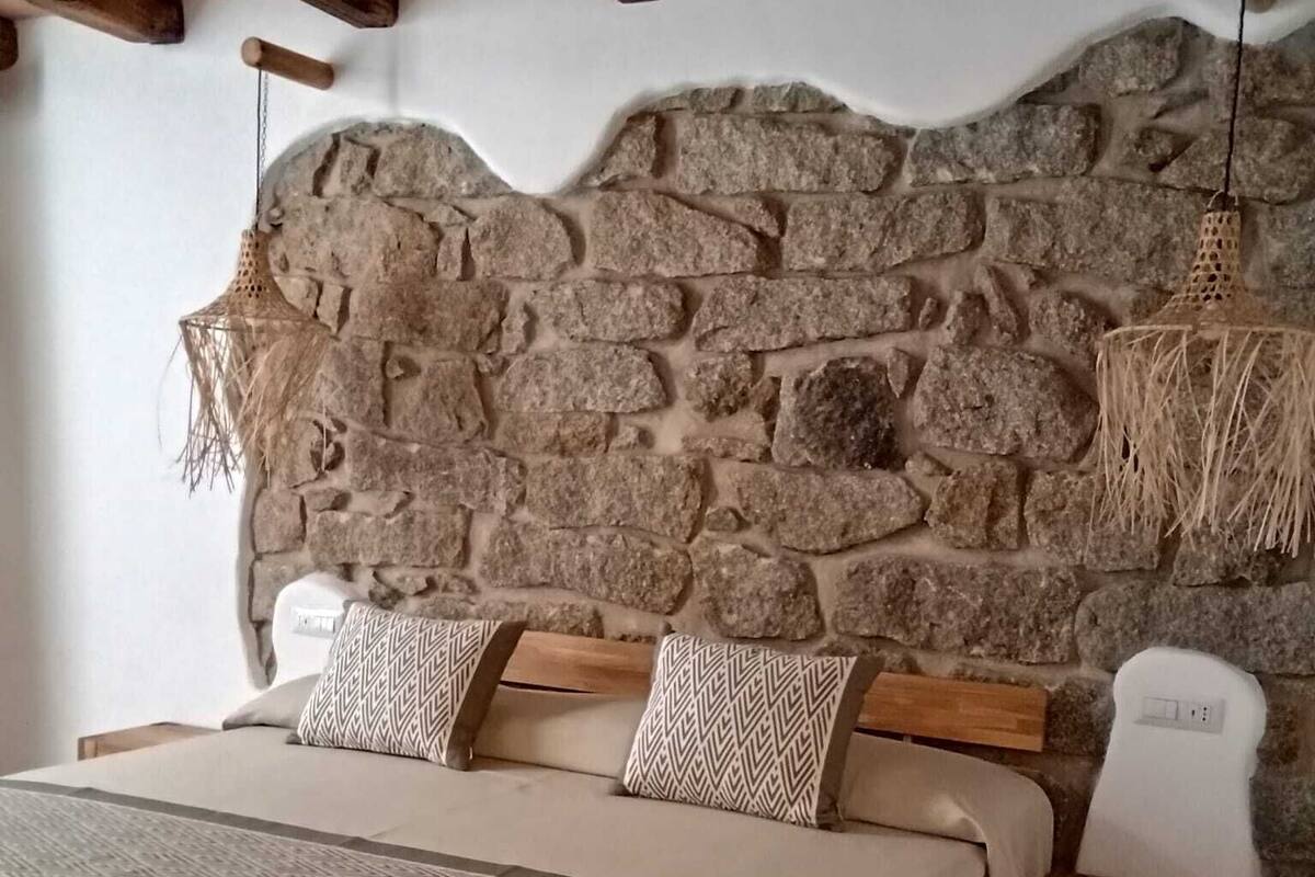 Ein Schlafzimmer mit Steinwand-Bettvorleger, einem Bett mit gemusterten Kissen, einem Nachttisch und einer Pendelleuchte.