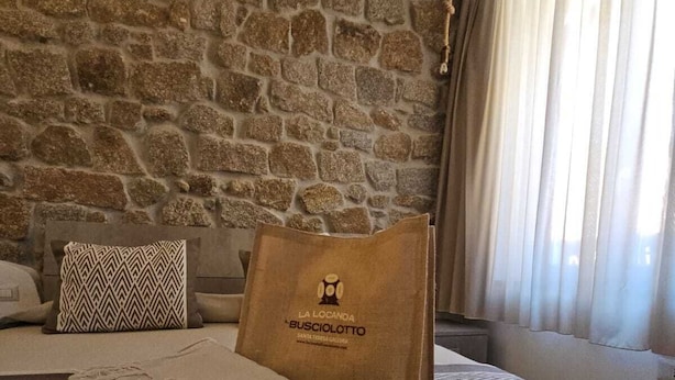 A cozy room with a stone wall, a bed with a patterned throw, a beige bag, and a window with curtains. - LOCANDA IL BUSCIOLOTTO (Santa Teresa di Gallura)