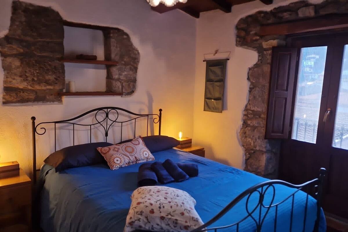 Un dormitorio con pared de piedra, una cama con estructura de metal, dos mesitas de noche con velas y una ventana con vistas.