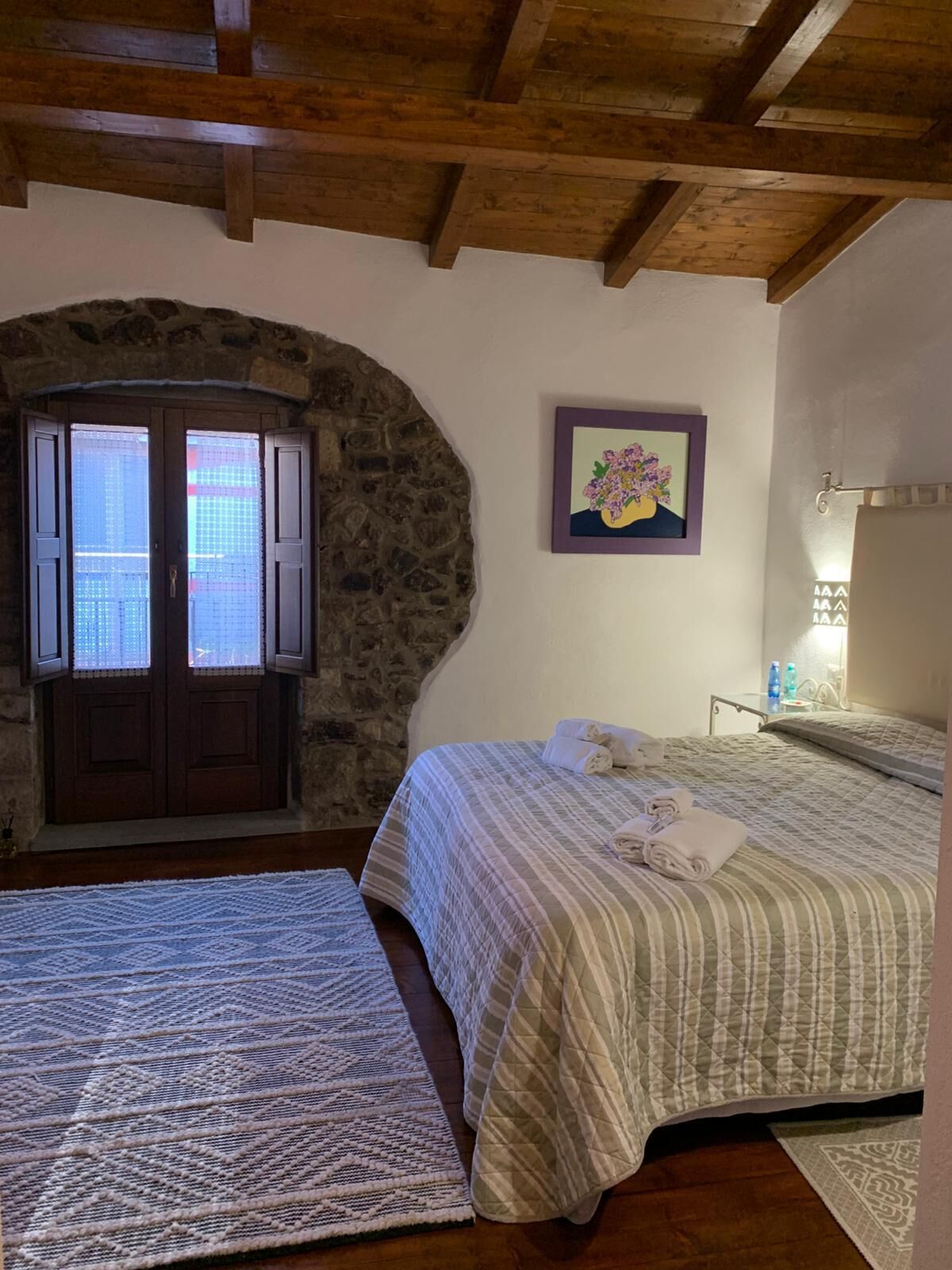Un dormitorio con un arco de piedra, una cama con colcha a rayas, un cuadro de flores y una mesita de noche con lámpara.