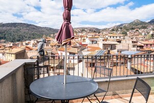 Un balcón con una mesa redonda y sillas, con vistas a un pueblo densamente poblado de tejados de terracota y un telón de fondo montañoso.