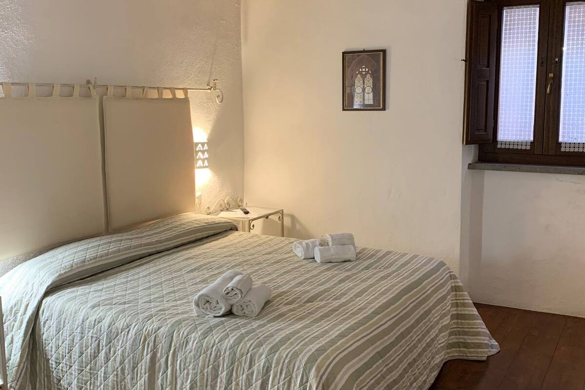 Un dormitorio con una cama grande, vigas de madera en el techo, una mesita de noche pequeña, una lámpara de noche y una ventana con cortinas. También se aprecia un cuadro enmarcado en la pared.