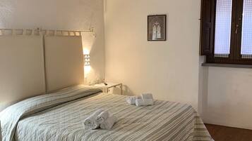 Un dormitorio con una cama grande, vigas de madera en el techo, una mesita de noche pequeña, una lámpara de noche y una ventana con cortinas. También se aprecia un cuadro enmarcado en la pared.