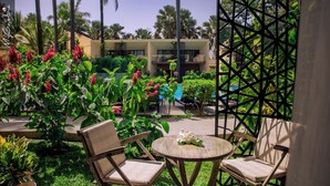 A patio with wicker chairs and a round table, surrounded by lush greenery and a pool. - Bakadaji Hotel (Serrekunda)
