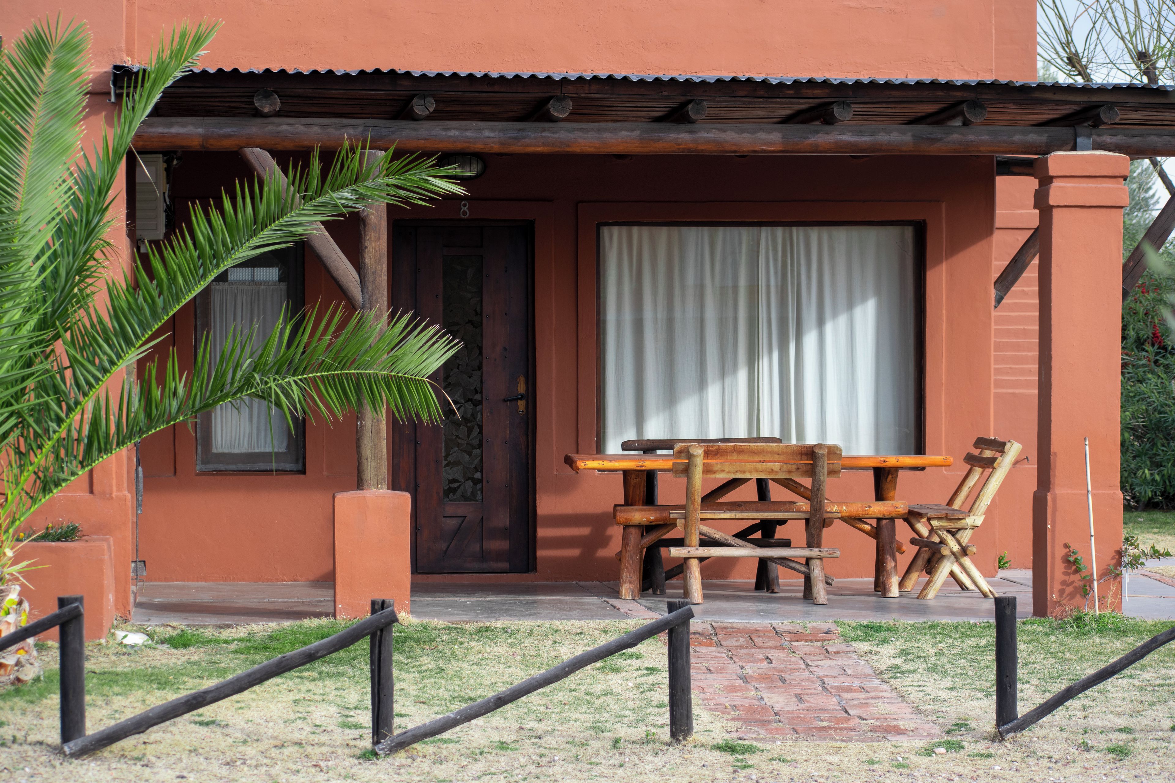 Mesa y sillas de madera frente a un edificio con puerta y ventana, rodeado de un pequeño jardín.
