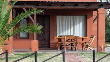 Mesa y sillas de madera frente a un edificio con puerta y ventana, rodeado de un pequeño jardín.