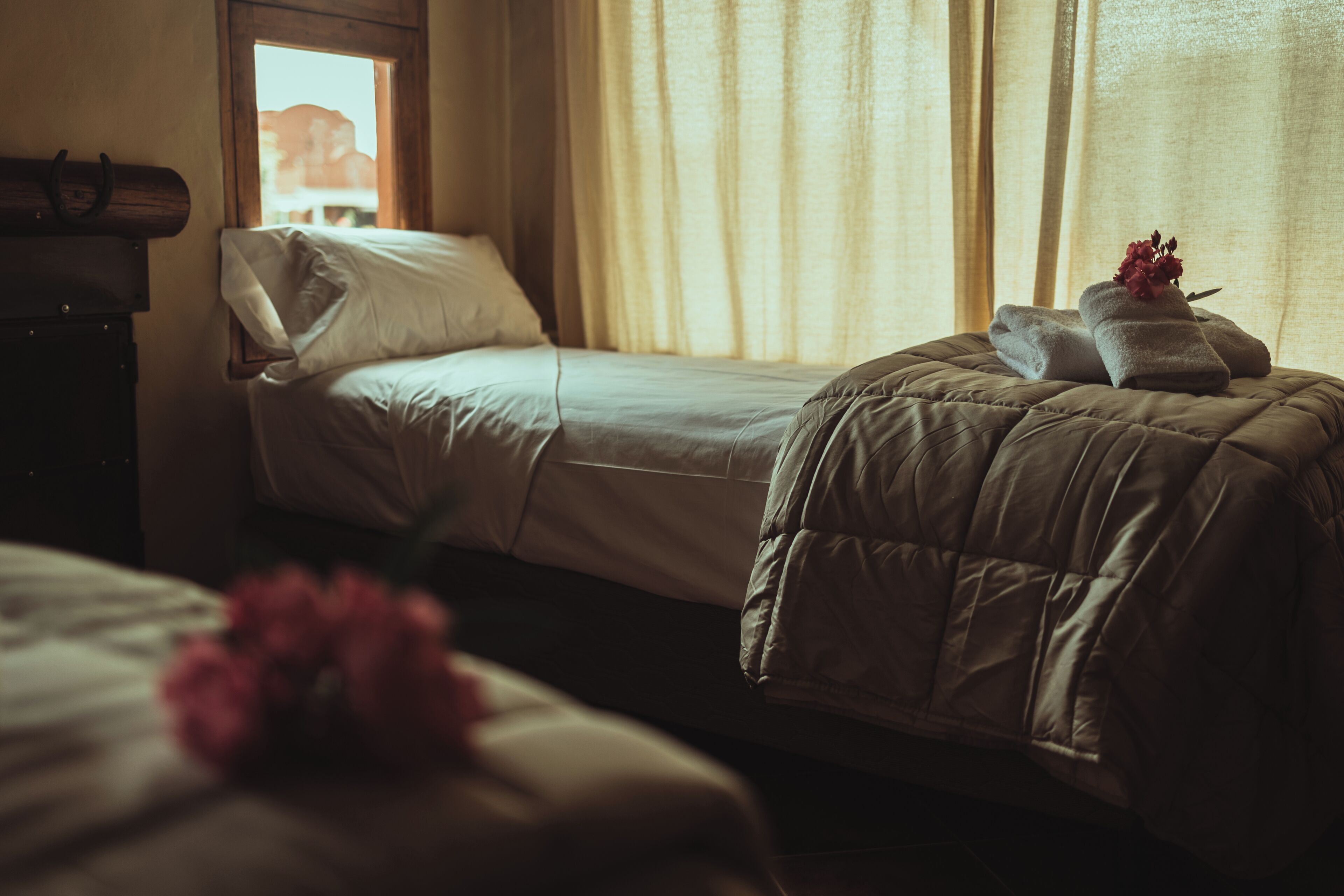 A bedroom with two beds, one with a quilted cover and the other with a plain cover. There is a window with curtains, a wooden headboard, and a dresser.