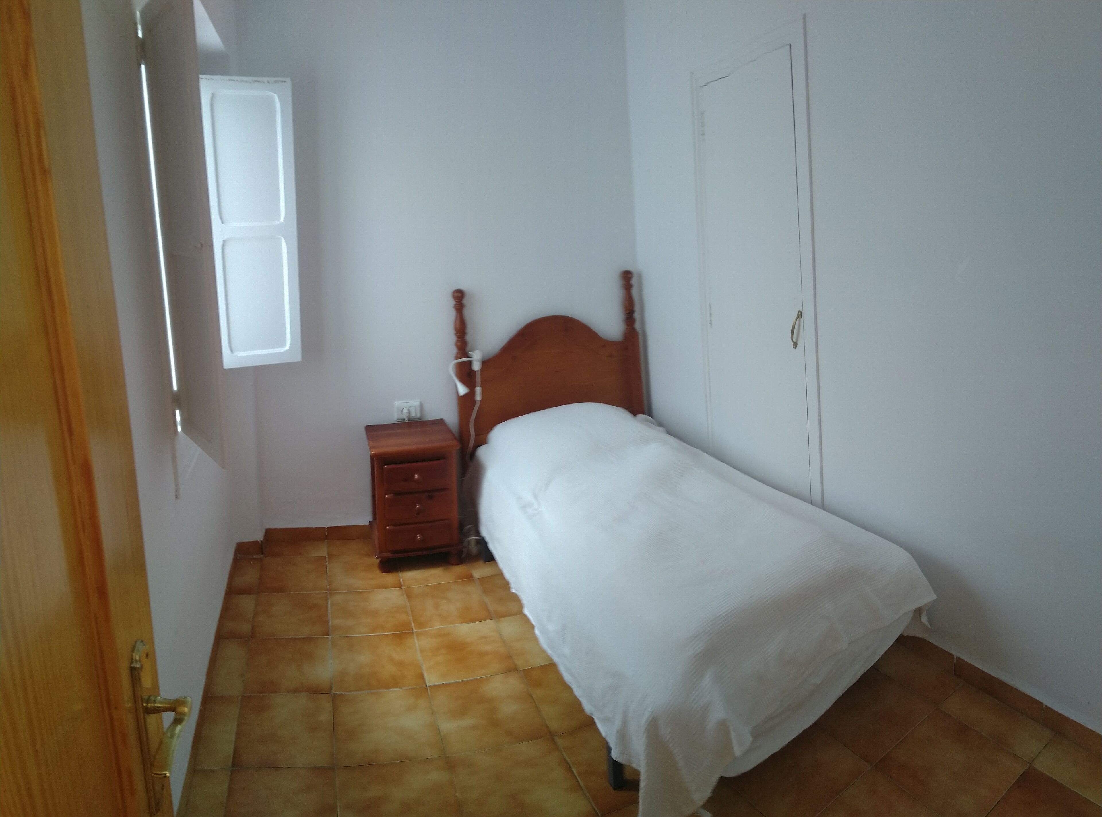 Quarto com cama de madeira, mesa de cabeceira, parede branca e janela.