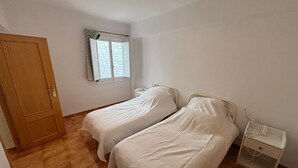 A room with two single beds, a wooden door, a window with patterned curtains, and a bedside table with a lamp. - Sunny beach apartment in Las Palmeres/Valencia (Sueca/Les Palmeres)