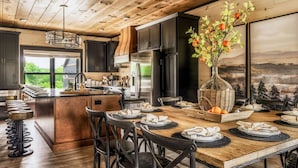 A modern kitchen with dark cabinets, a central island, and a dining area with chairs and a table set for a meal. - Roof Top Cabin~Water Park~Heater Indoor Pool~Games~PlayStation~Games~Mini-Golf~Views~Sleep 22 (Sevierville)