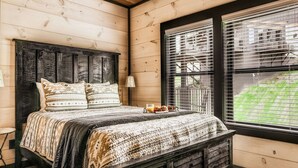 A wooden cabin bedroom with a bed, bedside table, lamp, and window with blinds. - Roof Top Cabin~Water Park~Heater Indoor Pool~Games~PlayStation~Games~Mini-Golf~Views~Sleep 22 (Sevierville)