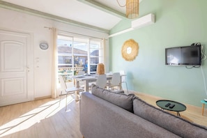 Quarto com sofá, mesa de jantar com cadeiras, TV fixada na parede e janela com vista para edifícios.