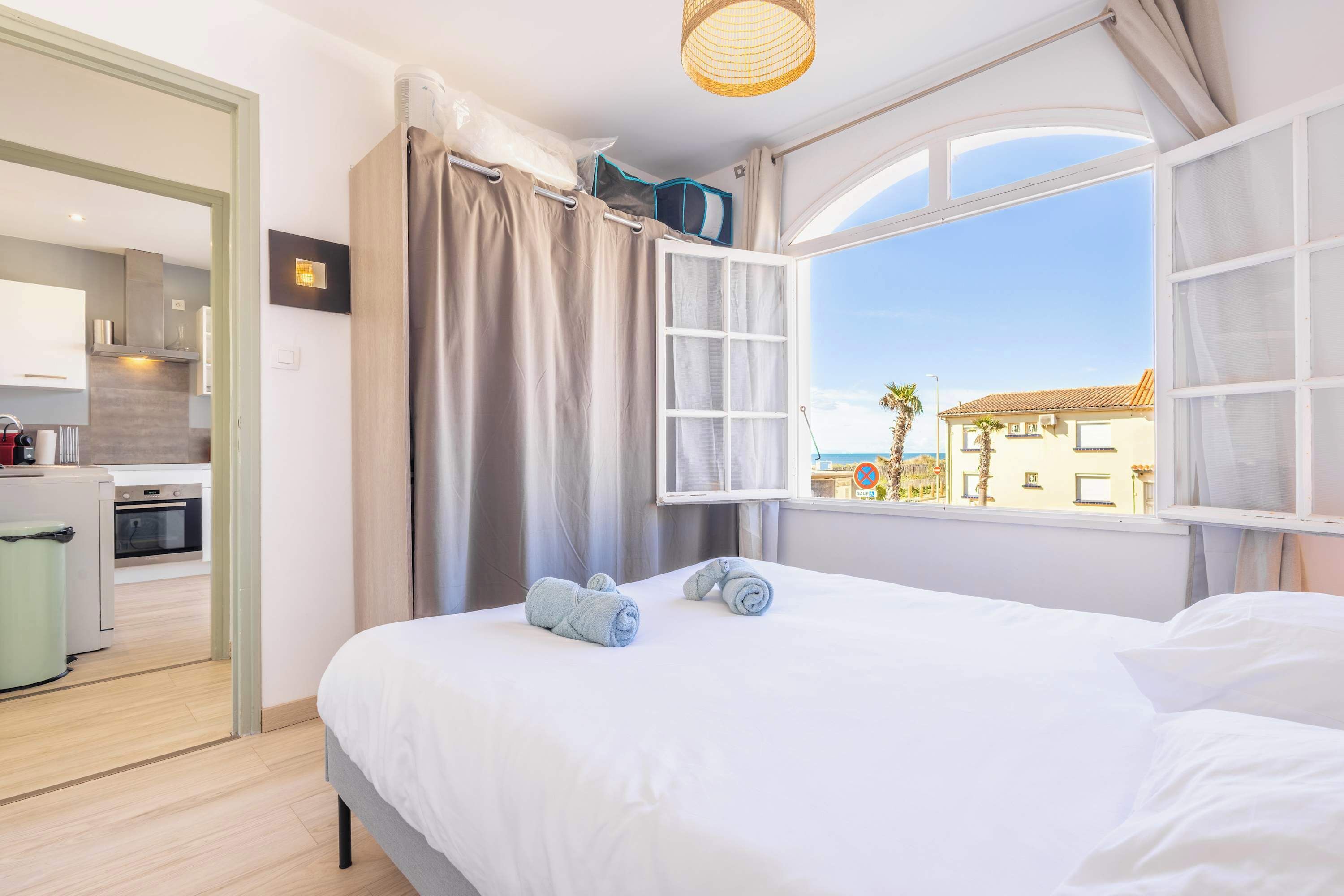 A bedroom with a bed, a window with curtains, a view of the outside, and a kitchenette in the background.