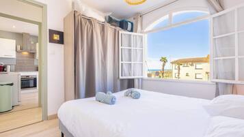 A bedroom with a bed, a window with curtains, a view of the outside, and a kitchenette in the background.