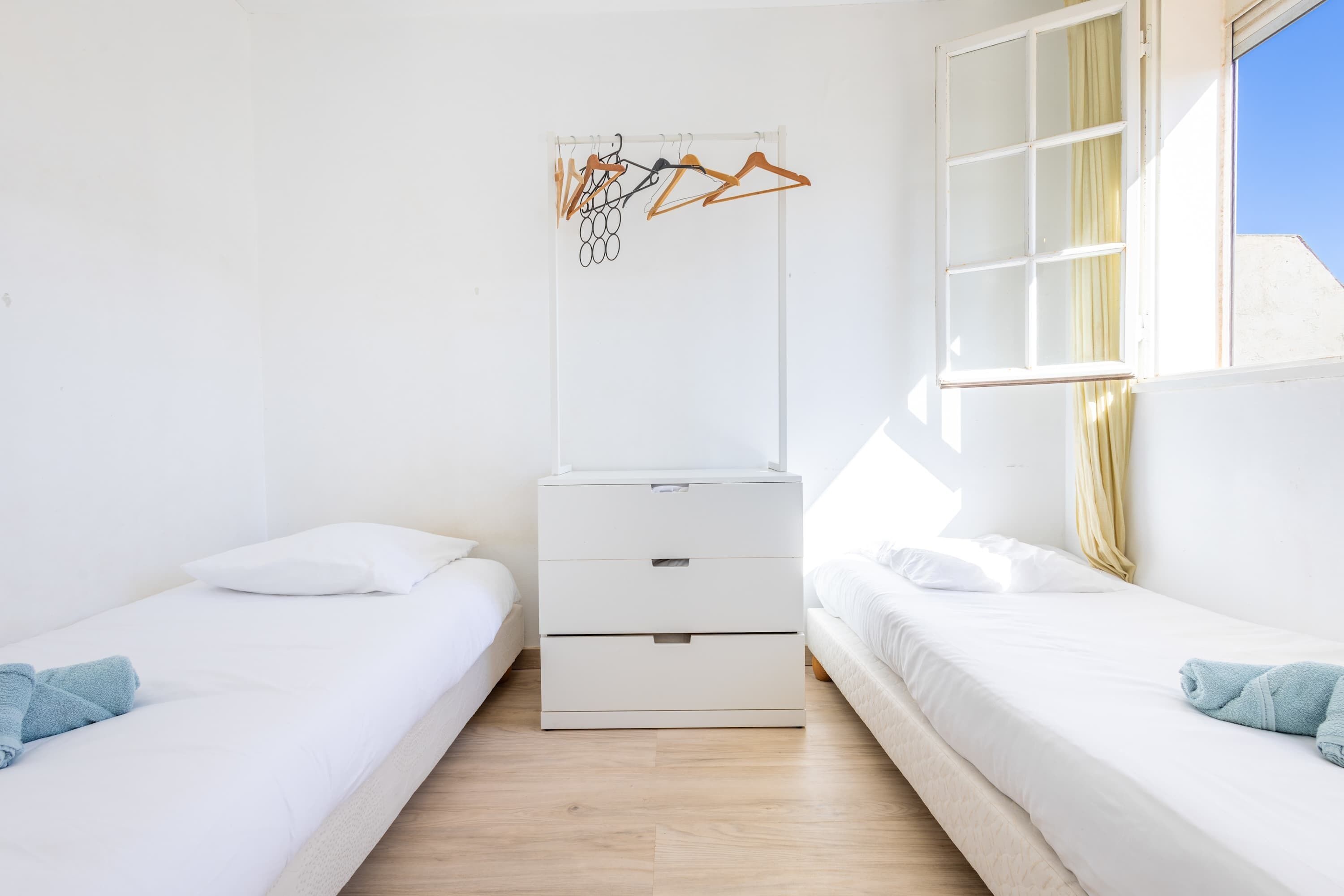 Een kamer met twee bedden, een witte ladekast, een raam met gordijnen en een wandrek met kledinghangers.