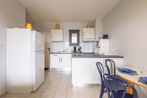 A compact kitchen with white cabinets, a refrigerator, a sink, and a dining area with chairs. - Cosy Apartment with Pool (Valras-Plage)