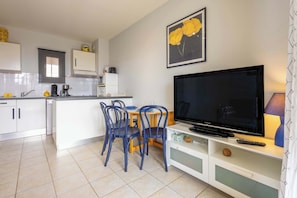 Une cuisine moderne avec des meubles blancs, un coin repas avec des chaises bleues, un téléviseur à écran plat et un tableau encadré au mur.