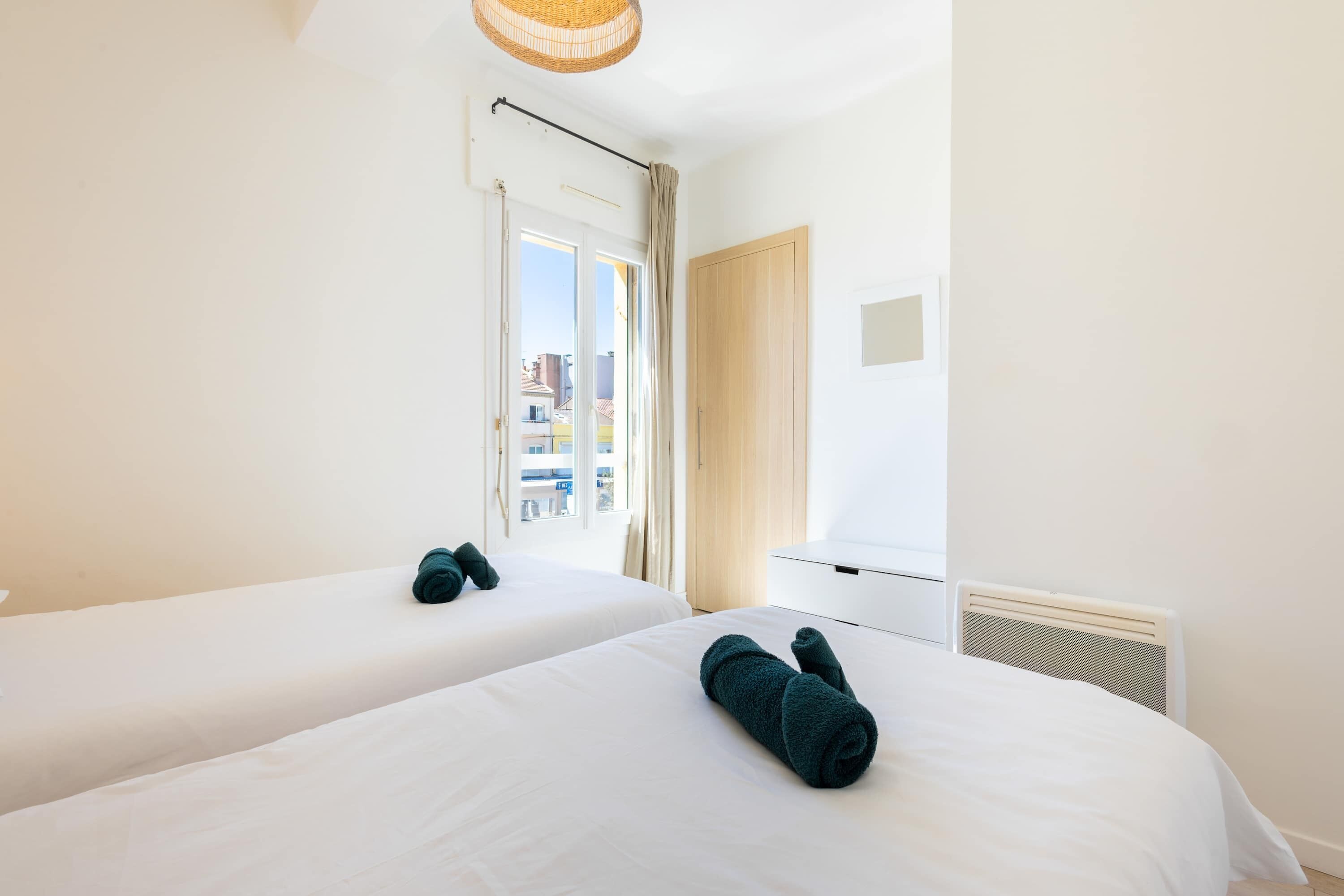 A hotel room with two beds, a window with curtains, a white radiator, and a wooden door.