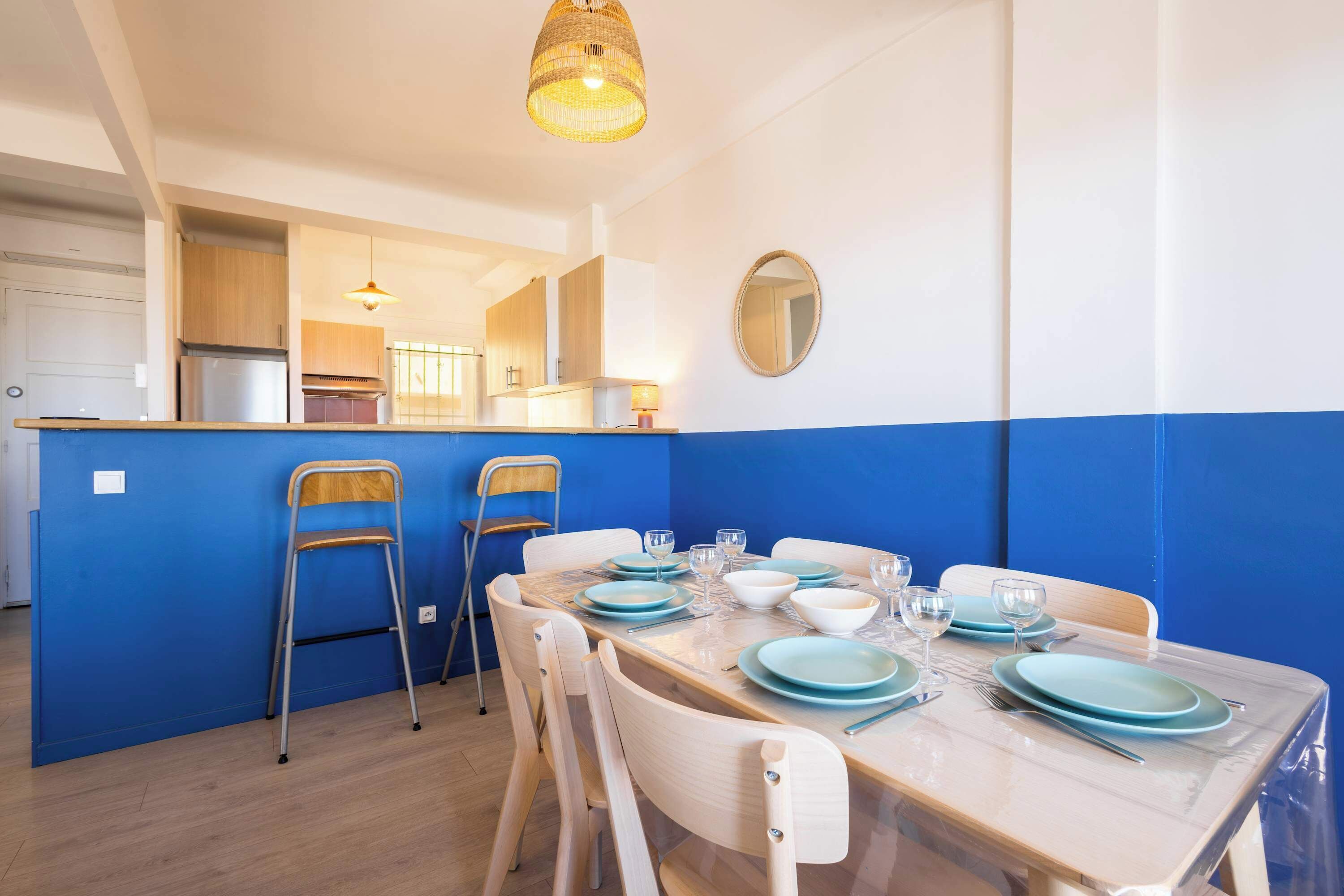 Comedor con una mesa preparada para cuatro personas, sillas, una cocina al fondo y una lámpara colgante.