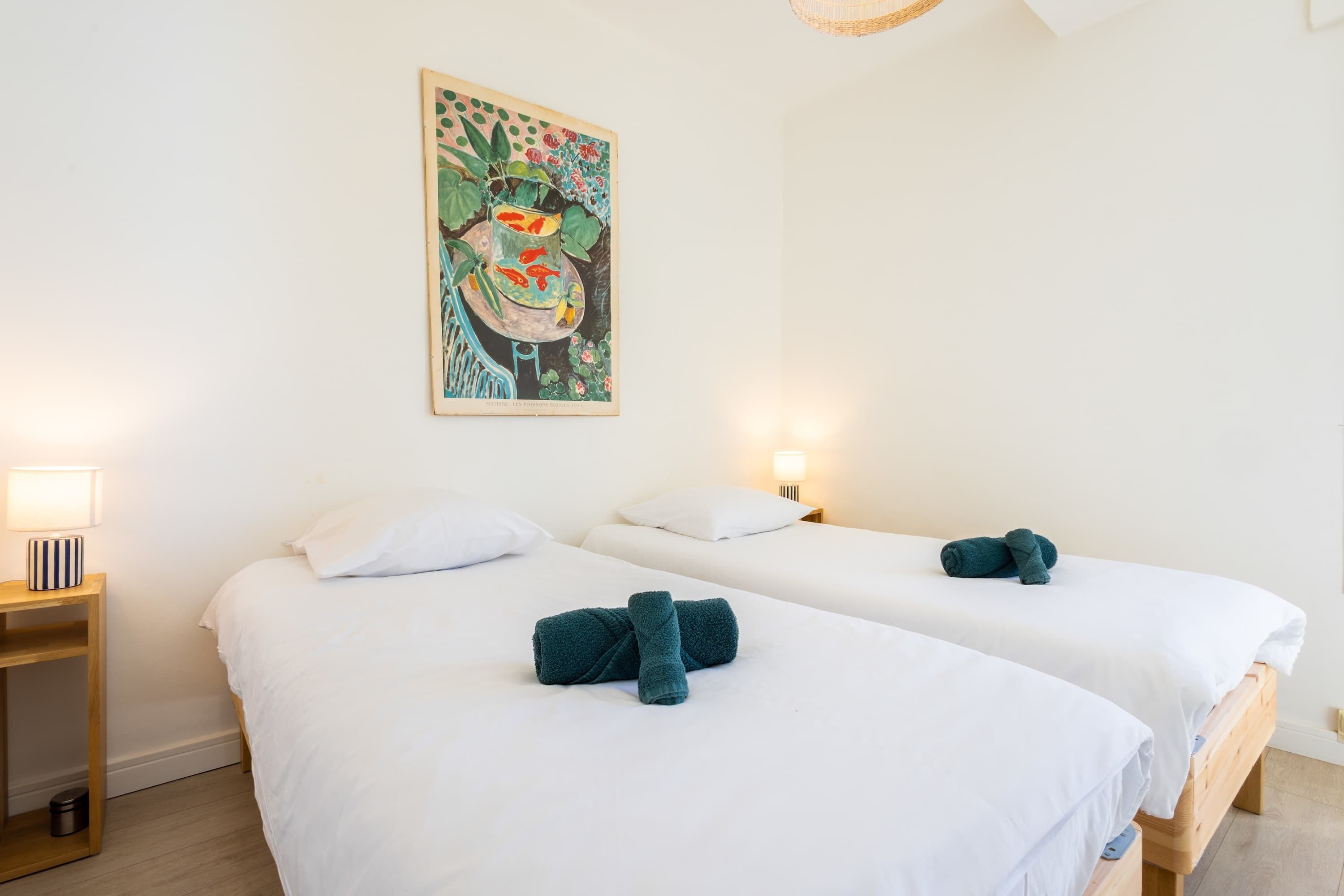 A hotel room with two beds, white linens, teal towels, a wall painting, bedside lamps, and a wooden floor.