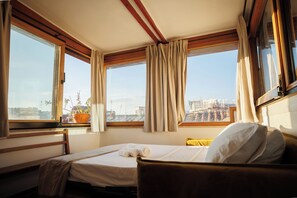 A hotel room with a bed, a window with curtains, a view of the city, and a potted plant on the windowsill.