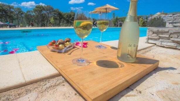 A wooden tray with a bottle of white wine, two wine glasses, and a bowl of assorted chocolates by a poolside.