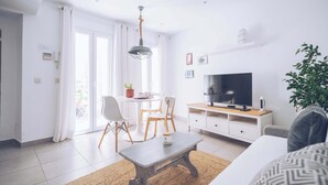 Ein modernes Wohnzimmer mit Sofa, Couchtisch und Fernseher. Es gibt einen Essbereich mit Stühlen und einem Tisch, sowie eine Pflanze in der Ecke.
