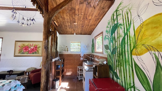 Un interior rústico con techo de madera, una zona de cocina con pileta y armarios, una mesa de comedor y un mural grande en la pared.