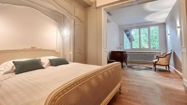 A bedroom with a large bed, a wooden chair, and a view of a window with greenery outside. - `Villa du Parc`*Parking*Proche centre*Calme (Montauban)