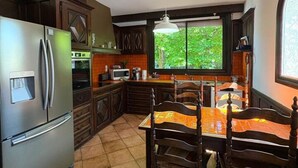 A kitchen with dark wooden cabinets, a stainless steel refrigerator, a dining table with chairs, and a window providing a view of greenery outside. - `Villa du Parc`*Parking*Proche centre*Calme (Montauban)
