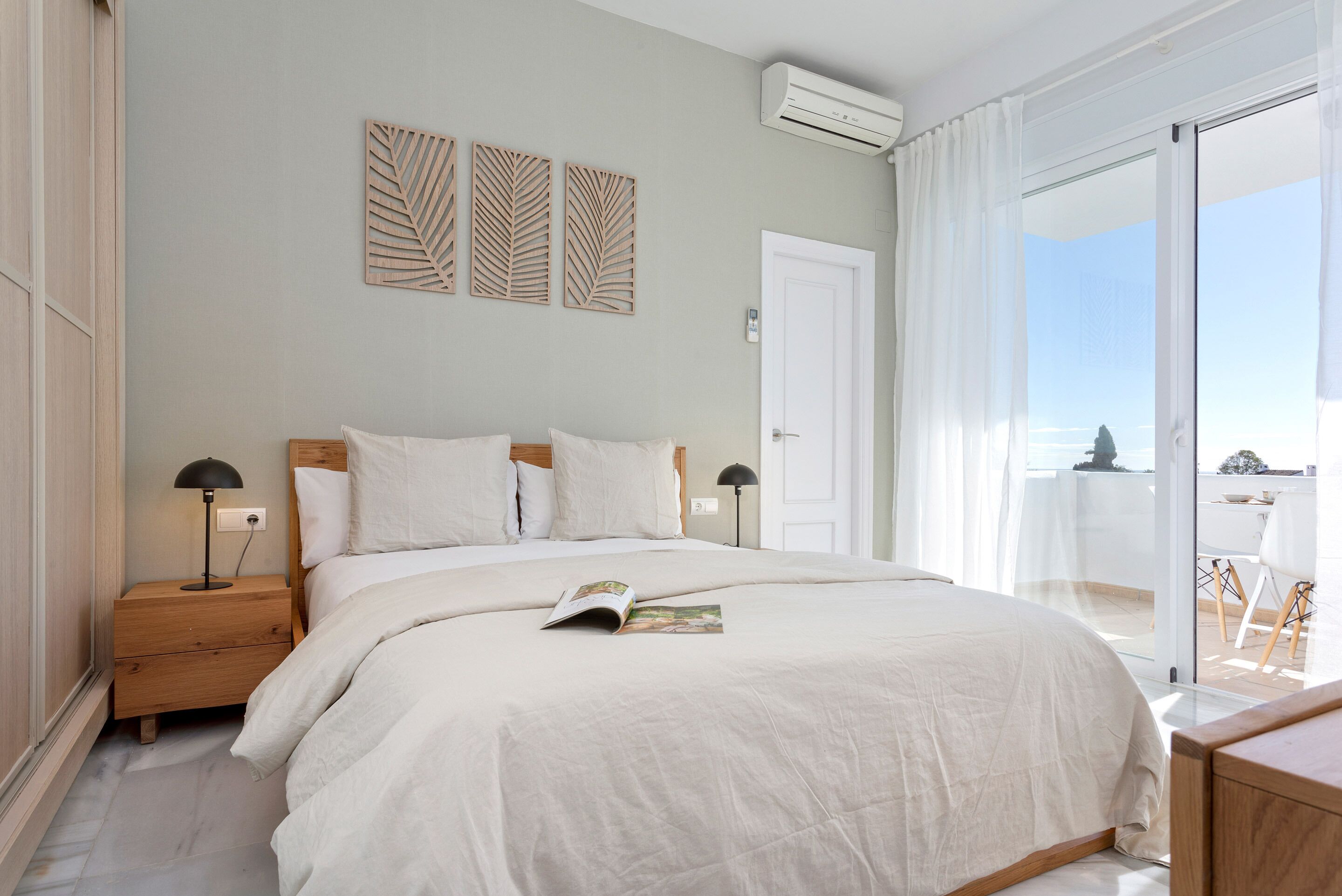 A bedroom with a large bed, a nightstand, a reading lamp, and a view of the outside.