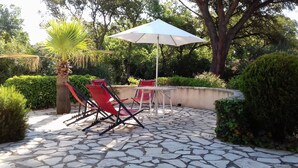 A patio area with stone flooring, red lounge chairs, a white umbrella, and lush greenery.
