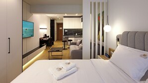 A modern hotel room with a neatly made bed, a desk with a laptop, a sofa, and a kitchen area in the background.