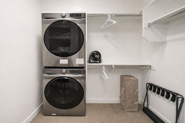 A laundry room with two stacked front-loading washing machines, a drying rack, a fan, and a bag of laundry.
