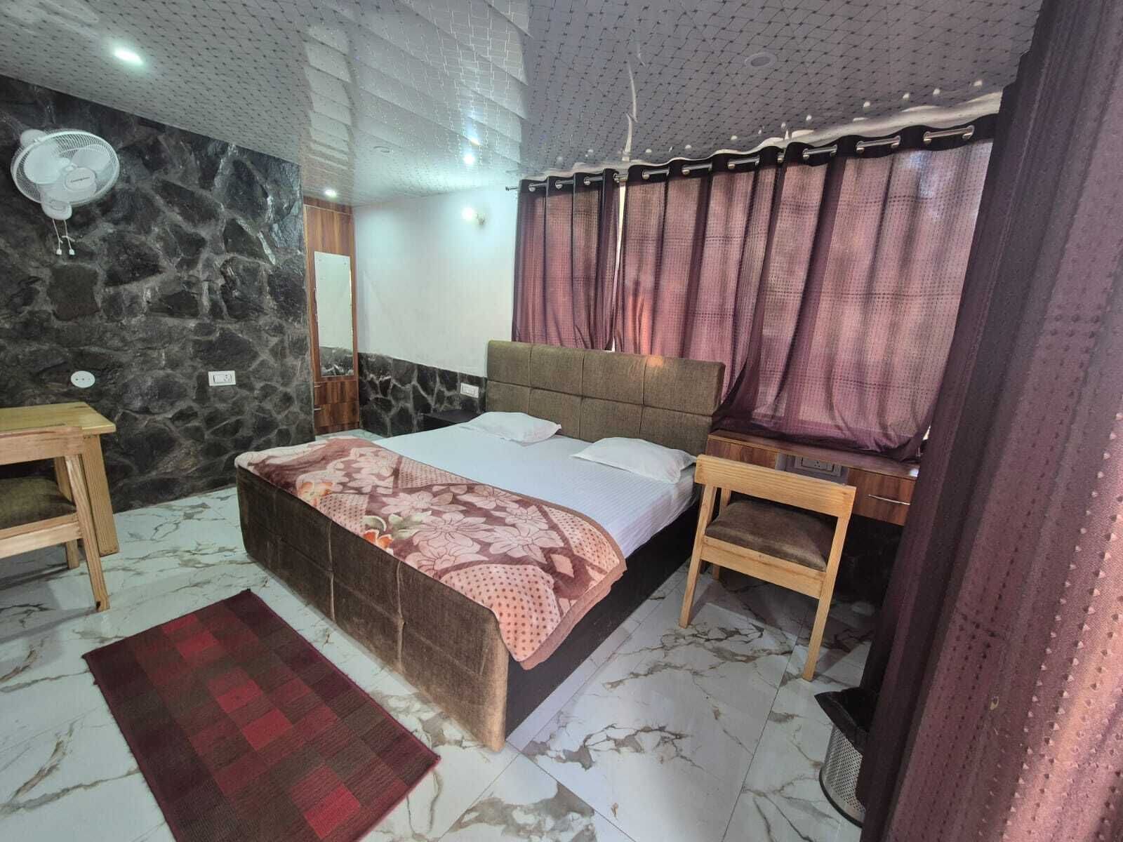 A bedroom with a bed, a nightstand, a wall-mounted fan, a mirror, and a red and brown patterned rug.