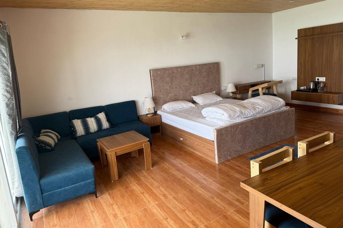 Egy modern hálószoba, melyben egy nagy ágy, egy kanapé, egy fa asztal és egy kis éjjeliszekrény található, az éjjeliszekrényen pedig egy lámpa van.