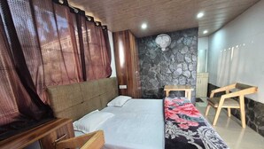 A bedroom with a bed, wooden nightstands, a desk, a chair, a fan, and a stone wall. - Echoes of the Himalayas (Suwakholi)