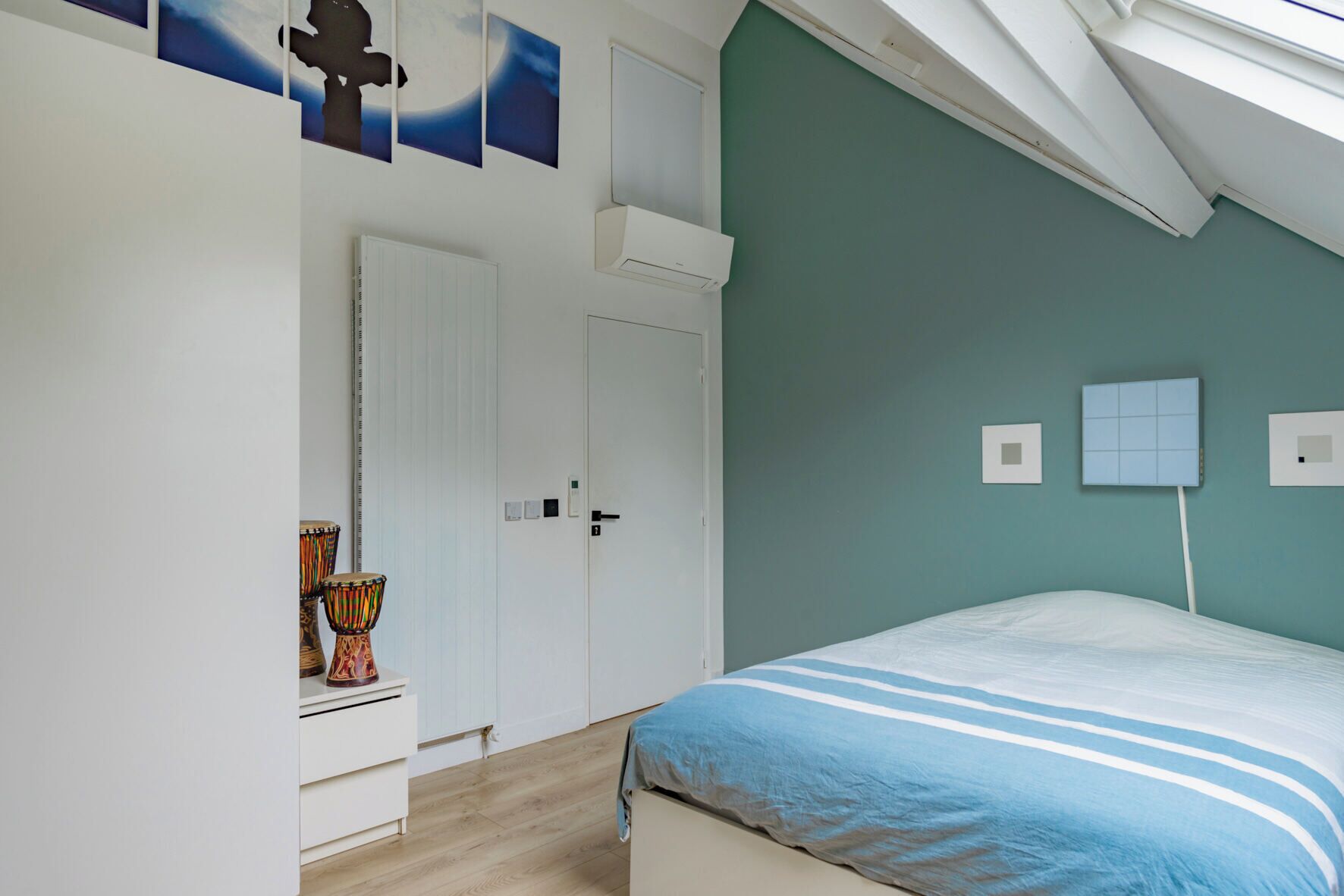 Un dormitorio con techo inclinado, una cama con cubrecama a rayas azules, un aparador blanco, un instrumento de percusión y una puerta con un número.