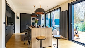 Cocina moderna con una isla central, taburetes de bar, iluminación colgante y vistas al exterior a través de amplias ventanas.
