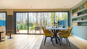 Un comedor moderno con una mesa grande y sillas, vista a través de puertas de vidrio a una terraza exterior, y un estante azul con objetos decorativos.