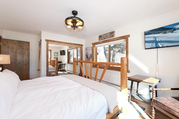 Un dormitorio con una cama grande, cabecera de madera, ventilador de techo, televisor y ventana con vistas a árboles y montañas.
