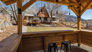 A wooden deck with two chairs and a table, overlooking a house with a balcony and a view of a mountainous landscape. - Private home on 5 acres - 5 min. from downtown Durango - Hot Tub/Game Room (Durango)