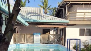 A pool area with a wooden deck, a sign indicating a pool service, and a building with a metal roof in the background. - Bluelight Village Hotel (Nadi)