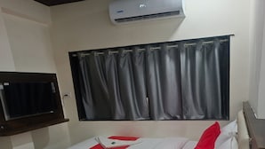 A single bed with a red and white bedspread, a red pillow, and a white pillow. There is a flat-screen TV mounted on the wall to the left of the bed. An air conditioning unit is mounted above the window, which has dark curtains. - HOTEL Sai Leela Residency (Mumbai)