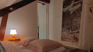 Et soveværelse med en seng, et natbord med en lampe, et billede på væggen og en dør, der fører til et andet rum.
