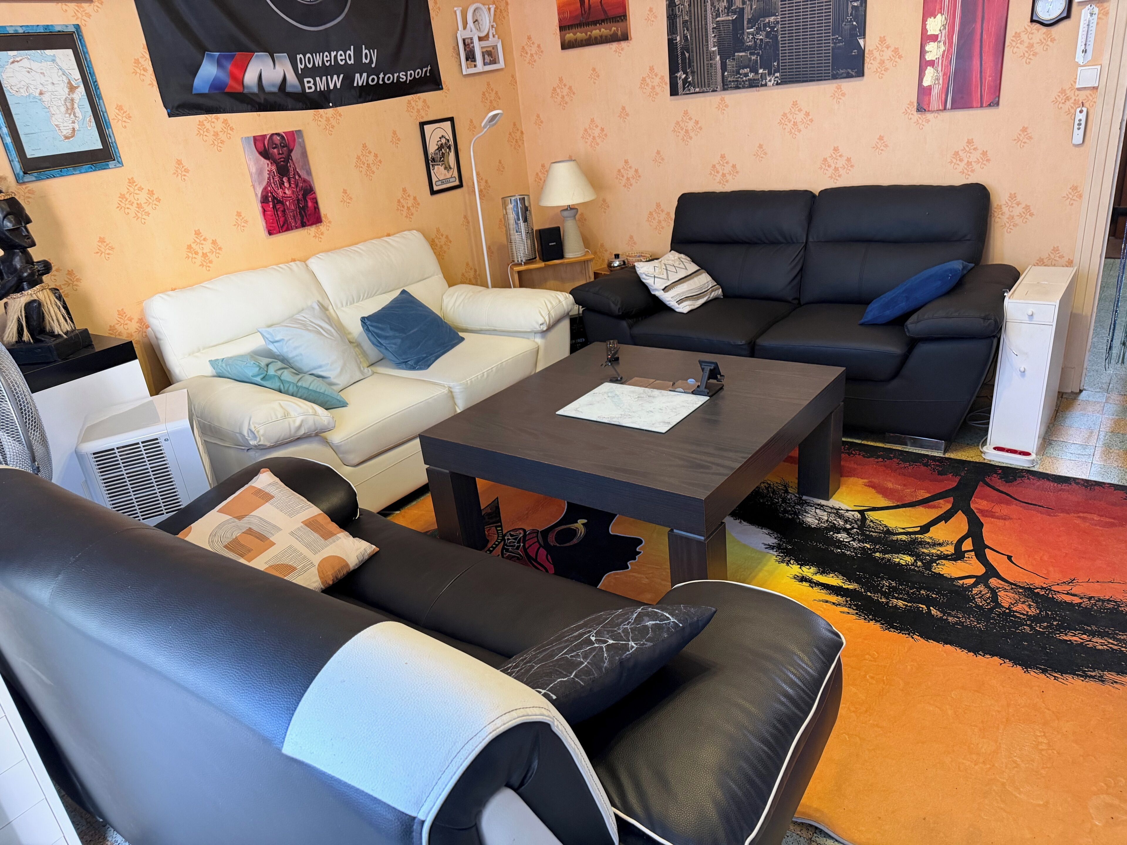 Een woonkamer met een witte bank, een zwarte bank en een salontafel. Er zijn wanddecoraties, een lamp en een klein wit kastje.