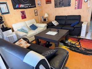 A living room with a white sofa, a black sofa, and a coffee table. There are wall decorations, a lamp, and a small white cabinet.