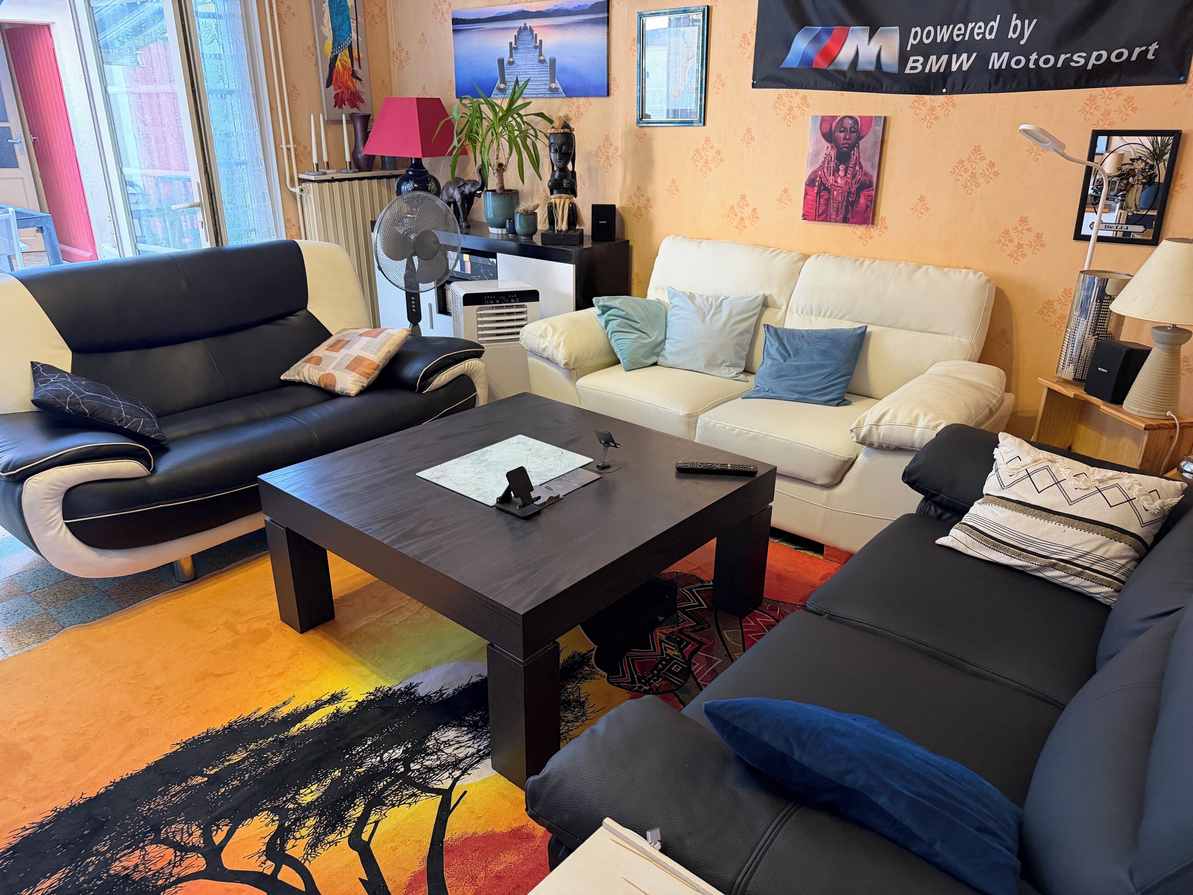Ein Wohnzimmer mit einer schwarzen Ledersofa, einem weißen Sofa und einem Couchtisch. Dort steht eine Lampe, eine Pflanze und ein gerahmtes Bild an der Wand.