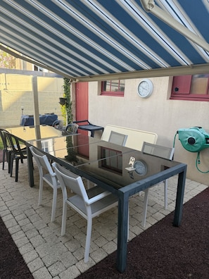 A covered patio area with a table and chairs, a clock on the wall, and a green hose on the side. - Bed and breakfast USA (Mayenne)